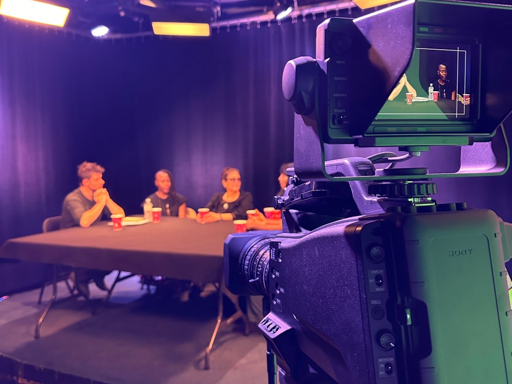 Camera filming a panel of four people seated at a table with a black backdrop.